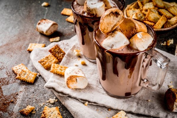 Traditional fall and winter cocktails, alcohol. Campfire hot chocolate eggnog cocktail with salted crackers and roasted marshmallow, in two mugs, on black stone table, copy space